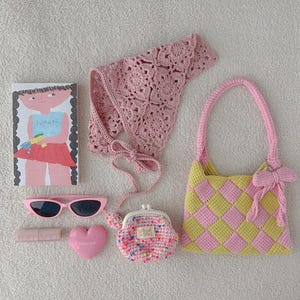 May include: A collection of pink accessories, including a crochet top, sunglasses, a heart-shaped item, a small coin purse, a lipstick tube, a book with a girl illustration, and a checkered handbag with a bow.