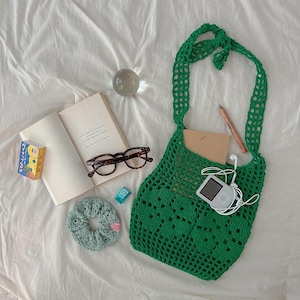 May include: A bright green crochet shoulder bag with an open mesh pattern. The bag contains a tan notebook, a white digital music player with white wired earbuds, and a wooden pencil. Nearby are a book, tortoise-shell glasses, a scrunchie, and a small box.