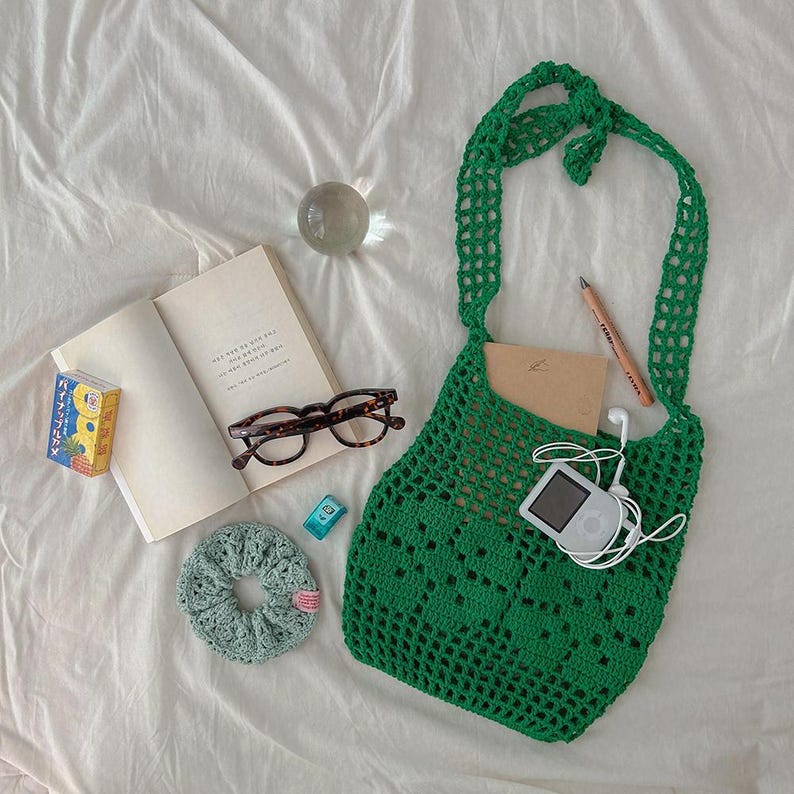 May include: A bright green crochet shoulder bag with an open mesh pattern. The bag contains a tan notebook, a white digital music player with white wired earbuds, and a wooden pencil. Nearby are a book, tortoise-shell glasses, a scrunchie, and a small box.