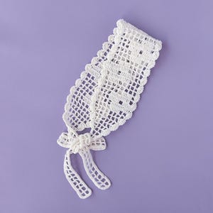 Puede incluir: Una diadema blanca de ganchillo hecha a mano con un delicado dise&ntilde;o calado. La diadema presenta un borde festoneado y un detalle de lazo en el centro, con lazos largos y colgantes. El art&iacute;culo se muestra sobre un fondo morado liso.