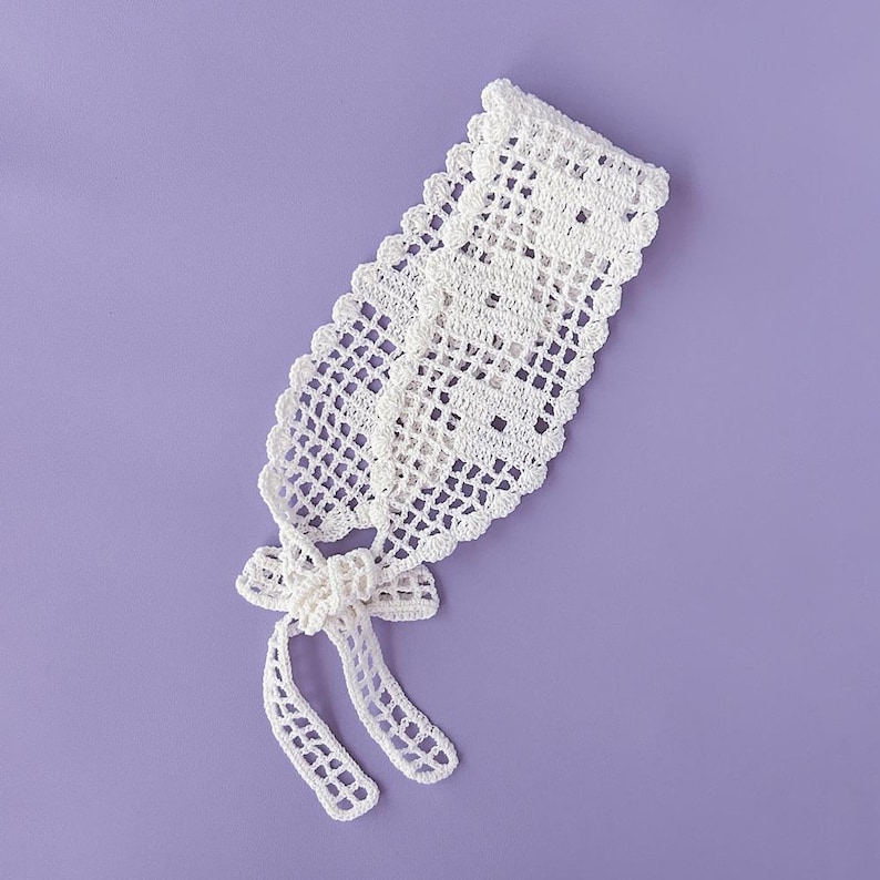 Puede incluir: Una diadema blanca de ganchillo hecha a mano con un delicado dise&ntilde;o calado. La diadema presenta un borde festoneado y un detalle de lazo en el centro, con lazos largos y colgantes. El art&iacute;culo se muestra sobre un fondo morado liso.