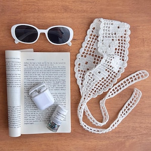 Puede incluir: Toma cenital de una colecci&oacute;n de accesorios sobre una superficie de madera. Incluye gafas de sol blancas, una diadema de ganchillo blanca, un libro, un par de auriculares inal&aacute;mbricos blancos y un frasco de esmalte de u&ntilde;as.