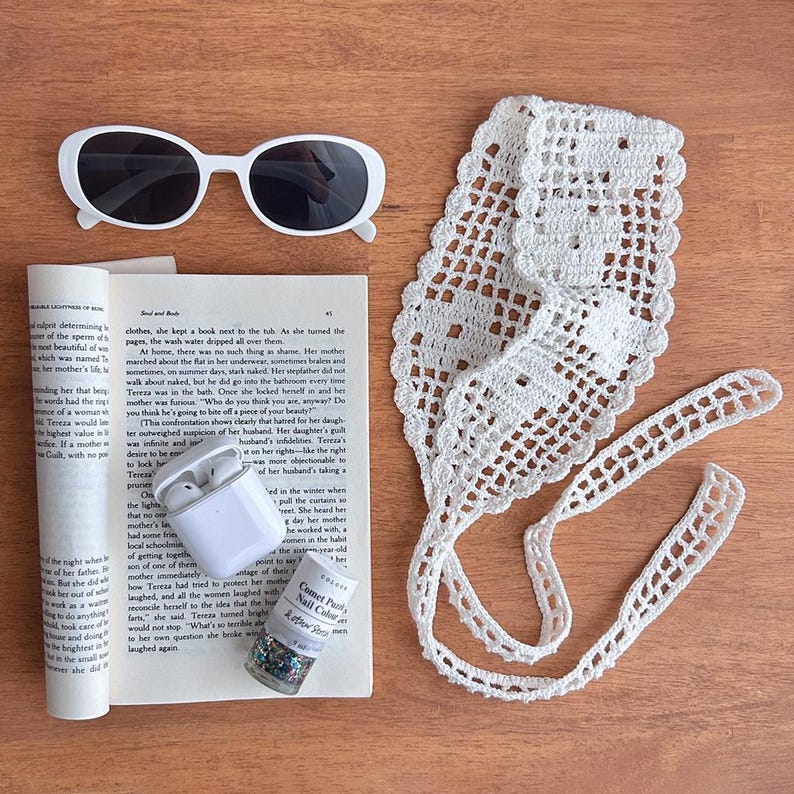 Puede incluir: Toma cenital de una colecci&oacute;n de accesorios sobre una superficie de madera. Incluye gafas de sol blancas, una diadema de ganchillo blanca, un libro, un par de auriculares inal&aacute;mbricos blancos y un frasco de esmalte de u&ntilde;as.