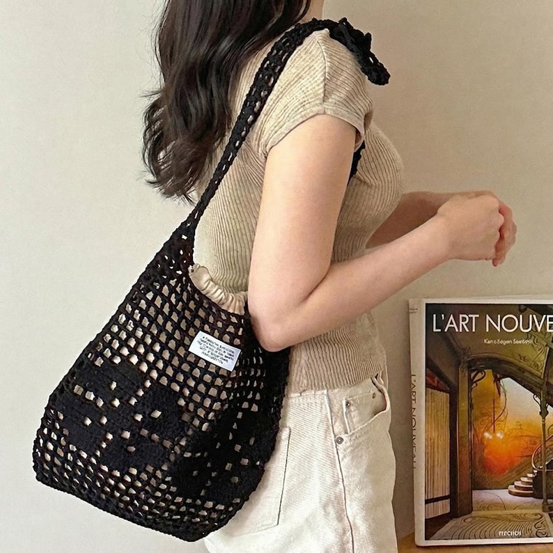 May include: A black crochet shoulder bag featuring an open-knit grid pattern and a small white rectangular label with black text. The bag has a drawstring closure and a long strap, showcasing a textured, handmade appearance with a soft inner lining.