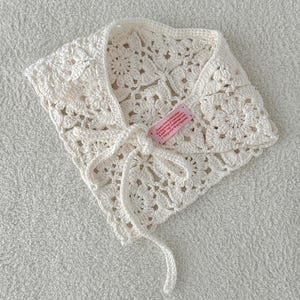 Könnte beinhalten: Cremefarbener Häkelschal mit floralem Muster und Bindverschluss. Der Schal hat ein zartes, offenes Design und ein kleines rosa Etikett mit Text. Der handgefertigte Schal ist ein stilvolles Accessoire.