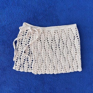 Puede incluir: Una falda cruzada de ganchillo en color crema que presenta un intrincado patrón calado con un dobladillo festoneado. La prenda incluye un cierre de lazo de ganchillo a juego en el lado izquierdo, lo que permite un ajuste regulable en la cintura.
