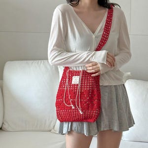 May include: A bright red crochet shoulder bag featuring an open-knit mesh pattern, a long matching strap, and a white drawstring closure. A small rectangular white fabric label with black text is stitched onto the front of the bag.