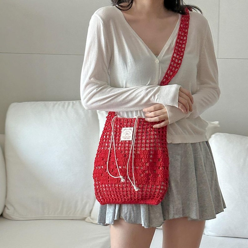 May include: A bright red crochet shoulder bag featuring an open-knit mesh pattern, a long matching strap, and a white drawstring closure. A small rectangular white fabric label with black text is stitched onto the front of the bag.