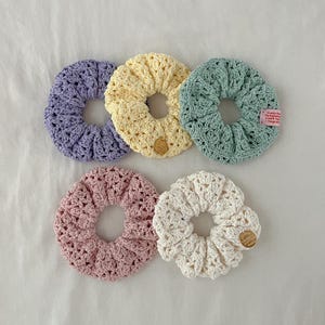 Può includere: Cinque scrunchies all'uncinetto fatti a mano in colori pastello: lavanda, giallo, verde menta, rosa e bianco. Ogni scrunchie presenta un design circolare con un foro centrale. Lo scrunchie verde menta ha una piccola etichetta con testo.