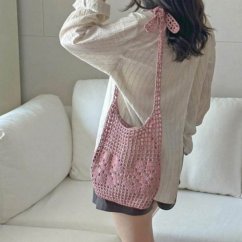 May include: A dusty pink crochet shoulder bag featuring an open-knit square pattern and a long strap that ties in a knot at the top. The bag is constructed from yarn with a visible texture and has a soft, slouchy shape that hangs from the shoulder.