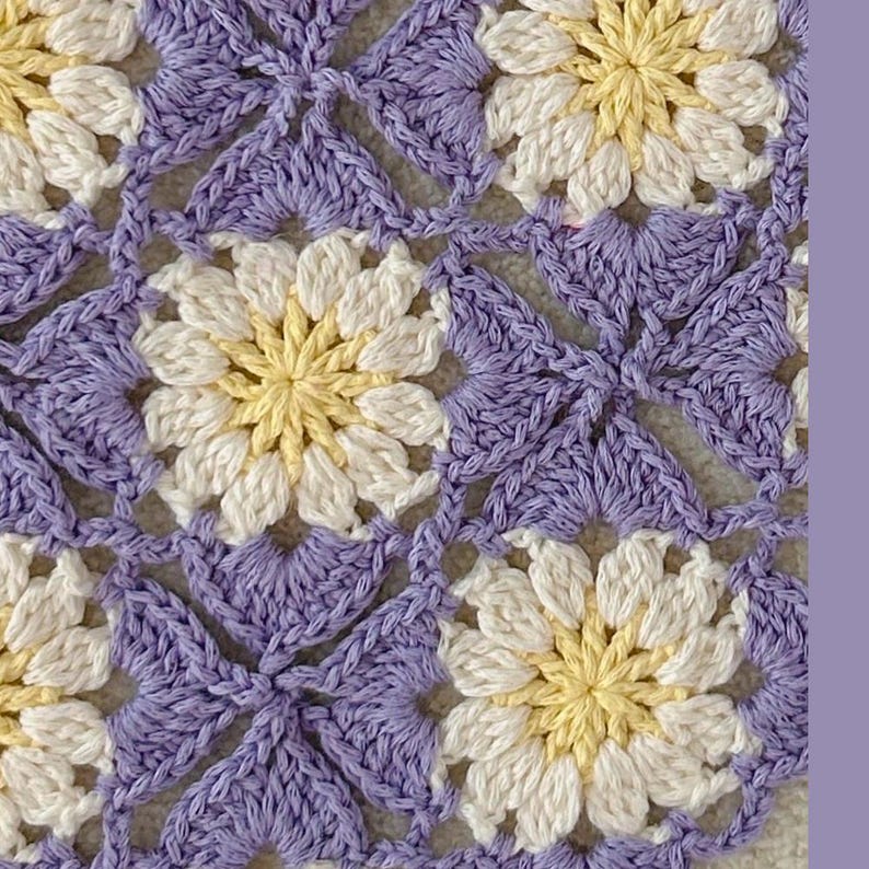 May include: Close-up of a crocheted floral pattern. The design features daisy-like flowers in white and yellow, surrounded by a lavender background. The crochet stitches create a textured, handmade appearance.