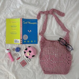 May include: A dusty rose crochet market bag with a tie strap, a blue book titled Hoot by Carl Hiaasen, a pink Hello Kitty Pez dispenser, a small pink and black crochet coin purse, a square pink compact, two coins, a wrapped candy, and black glasses.