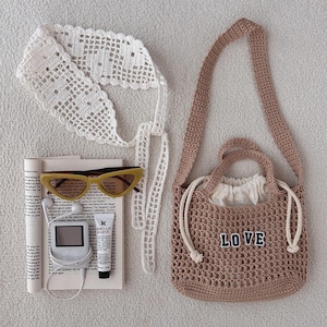Puede incluir: Una colecci&oacute;n de accesorios que incluye un bolso de crochet con "LOVE" cosido en la parte delantera, una diadema de crochet blanca, gafas de sol amarillas, un peque&ntilde;o reproductor de m&uacute;sica y b&aacute;lsamo labial. Los art&iacute;culos est&aacute;n colocados sobre un libro.