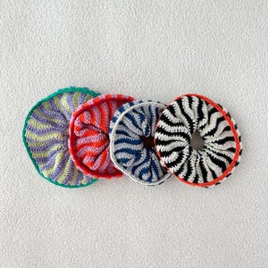 Puede incluir: Cuatro coleteros de ganchillo coloridos en fila. Los coleteros son en tonos morado, naranja, azul y blanco y negro. Cada coletero tiene un patrón y una combinación de colores únicos, con un diseño circular. Los coleteros están hechos de hilo.