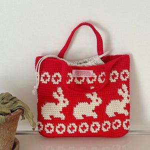 May include: Red crocheted tote bag with white bunny and flower designs. The bag has a white fabric lining and a pink label that reads "Crocheted Patchwork Bag - The beginning of a new story - a warm bag, handmade with love - a thing full of my happiness".