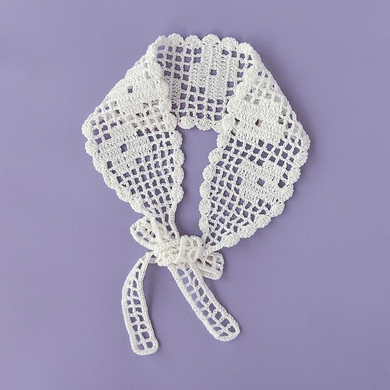 Puede incluir: Un cuello blanco de ganchillo con un borde festoneado y un cierre de lazo. El cuello presenta un delicado patr&oacute;n de tejido abierto y se muestra sobre un fondo lavanda. Un accesorio hecho a mano.