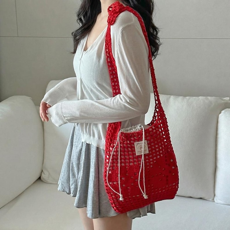 May include: A bright red crochet shoulder bag featuring an open-knit mesh pattern, a long strap, and a white drawstring closure at the top. A small rectangular white fabric label with black text is stitched onto the front of the red bag.