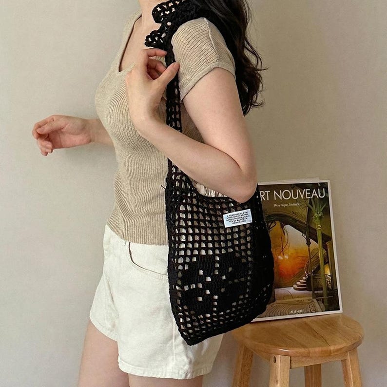 May include: A black crochet tote bag with an open-knit grid pattern and a small white rectangular label featuring black text. The bag has a single shoulder strap and is constructed from yarn, showcasing a textured, handmade appearance.