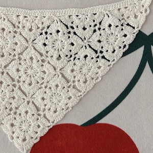 May include: Close-up of a white, crocheted shawl with a floral pattern. The shawl is triangular, with a delicate, openwork design. The crochet work is detailed, showcasing the texture and craftsmanship. The background is a patterned surface with a red cherry.