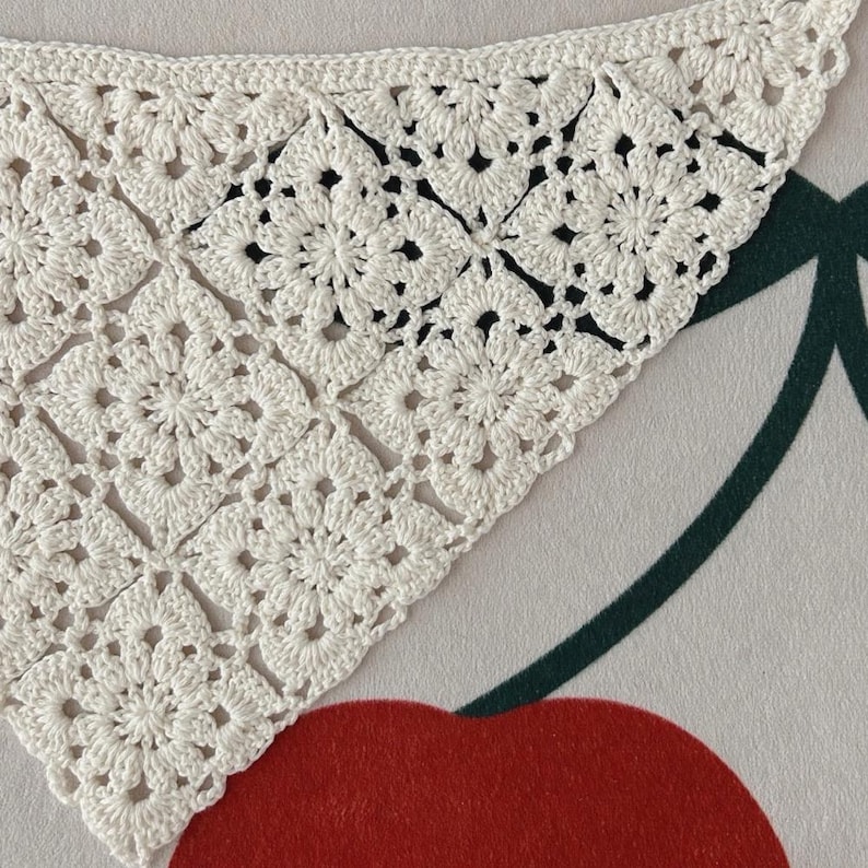 May include: Close-up of a white, crocheted shawl with a floral pattern. The shawl is triangular, with a delicate, openwork design. The crochet work is detailed, showcasing the texture and craftsmanship. The background is a patterned surface with a red cherry.