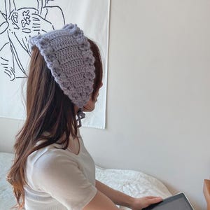 Puede incluir: Una mujer lleva una boina de ganchillo de color lila claro con un lazo atado. La boina tiene un patrón texturizado y se lleva sobre su largo cabello castaño.