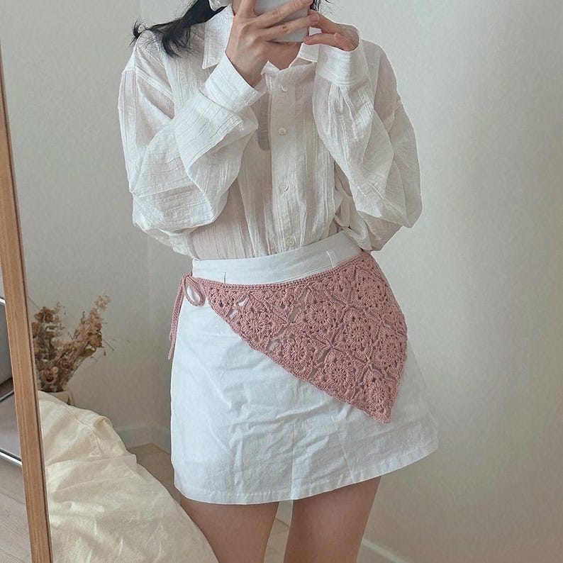 May include: A white skirt with a pink crochet overlay. The overlay is triangular and tied at the waist. The person is wearing a white button-down shirt.