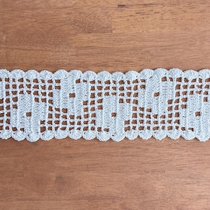 Puede incluir: Ribete de encaje de ganchillo blanco con borde festoneado. El dise&ntilde;o calado presenta un patr&oacute;n repetitivo de peque&ntilde;os cuadrados y elementos decorativos. El ribete se muestra sobre una superficie de madera.