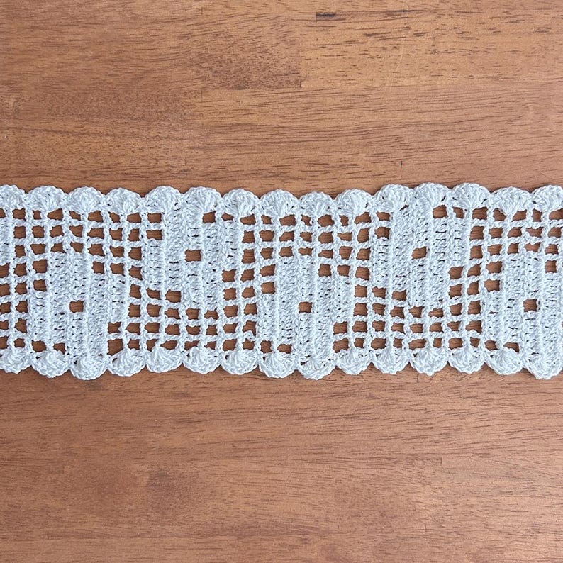 Puede incluir: Ribete de encaje de ganchillo blanco con borde festoneado. El dise&ntilde;o calado presenta un patr&oacute;n repetitivo de peque&ntilde;os cuadrados y elementos decorativos. El ribete se muestra sobre una superficie de madera.
