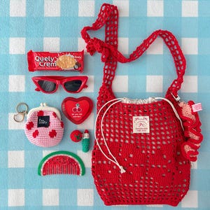 May include: A red crochet drawstring bag with a white label reading Handmade with love, a red Quely Crem snack pack, red cat-eye sunglasses, a pink and red crochet coin purse, a heart-shaped cherry compact, a small tomato charm, and a watermelon comb.