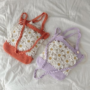 May include: Two handmade crochet tote bags. One is orange and white, the other is lavender and white. Both bags feature a daisy flower pattern. The bags have drawstring closures and braided straps. The bags are on a white sheet.