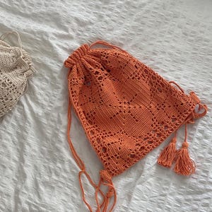 May include: An orange crochet drawstring bag with a geometric pattern. The bag has long drawstrings and decorative tassels. The bag is made of a soft, open-weave material, perfect for carrying items.