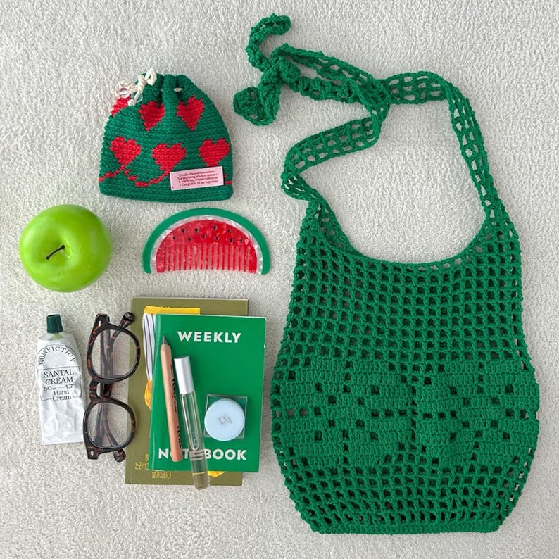 May include: A green crochet net bag, a green drawstring pouch with red hearts and a pink label, a green apple, a watermelon-shaped comb, a weekly notebook, a tube of hand cream, brown-rimmed glasses, a pencil, a clear pen, and a small round container.