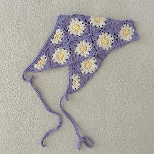 May include: A crocheted lavender head covering featuring a daisy pattern in white and yellow. The head covering has two long lavender ties. The item is handmade.