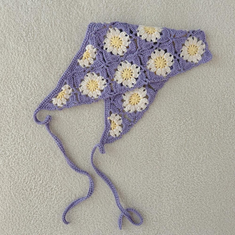 May include: A crocheted lavender head covering featuring a daisy pattern in white and yellow. The head covering has two long lavender ties. The item is handmade.