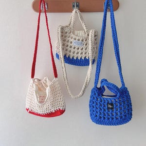 Könnte beinhalten: Drei handgefertigte Häkeltaschen in Rot, Creme und Blau hängen an einem Holzregal. Die rote Tasche ist eine kleine Umhängetasche mit Kordelzug. Die cremefarbene Tasche hat einen blauen Boden und einen kurzen Griff. Die blaue Tasche ist eine kleine Tragetasche.