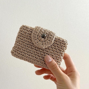 Può includere: Un porta carte beige all'uncinetto con chiusura a bottone. Il piccolo portafoglio è realizzato in un materiale intrecciato e strutturato. Il bottone è grigio scuro. Il porta carte è tenuto in una mano.