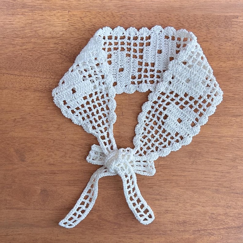 Puede incluir: Un cuello blanco de ganchillo con un delicado dise&ntilde;o calado. El cuello presenta un borde festoneado y un cierre de lazo. El accesorio hecho a mano se muestra sobre una superficie de madera.