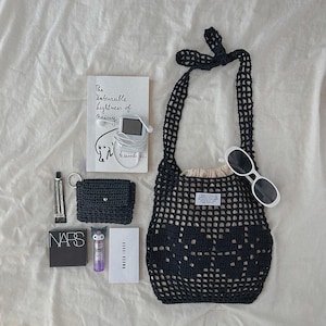 May include: A black crochet net bag with a beige drawstring pouch inside, a book titled The Unbearable Lightness of Being, a digital music player with white earbuds, a small black crochet wallet, white sunglasses, NARS compact, and Bobbi Brown box.
