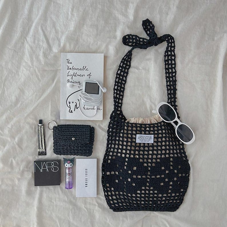 May include: A black crochet net bag with a beige drawstring pouch inside, a book titled The Unbearable Lightness of Being, a digital music player with white earbuds, a small black crochet wallet, white sunglasses, NARS compact, and Bobbi Brown box.