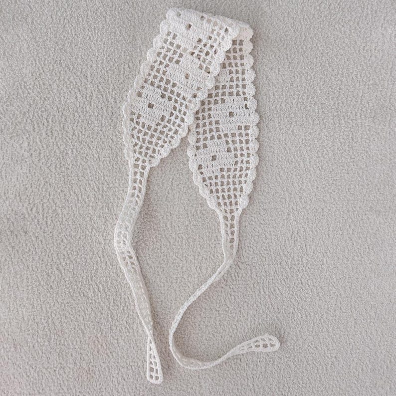 Puede incluir: Una diadema blanca de ganchillo de tejido abierto con un dise&ntilde;o delicado y calado. La diadema presenta una secci&oacute;n central rectangular con un patr&oacute;n repetido, que se estrecha en lazos largos y delgados. El art&iacute;culo se muestra sobre una superficie texturizada gris claro.
