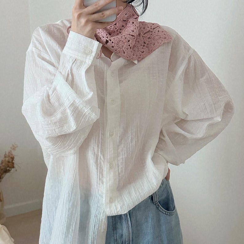 May include: A white button-down shirt with long sleeves and a pink crocheted collar. The shirt is paired with blue jeans. The collar is a delicate, openwork design. The shirt has a textured appearance.