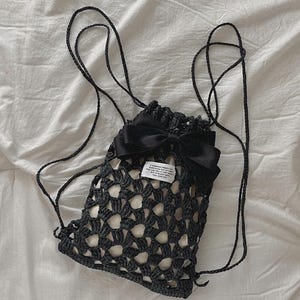 Puede incluir: Bolso negro de crochet con cordón, lazo negro grande y correas largas negras. El bolso tiene un interior blanco y una pequeña etiqueta con texto. El bolso está hecho de un material tejido de tejido abierto.