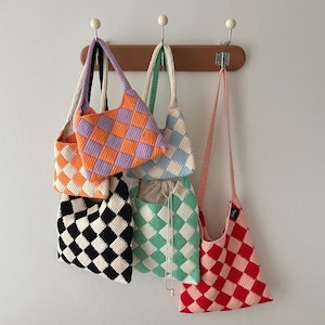 May include: A collection of six handmade crochet tote bags in various colors, including orange, purple, black, white, mint green, and red. Each bag features a checkered pattern and a long strap for shoulder or crossbody wear. The bags are hanging on a wooden rack.