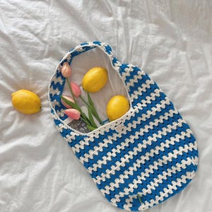 May include: A blue and white crochet tote bag with a striped pattern. The bag contains three yellow lemons and a few pink tulips. The bag is sitting on a white sheet.