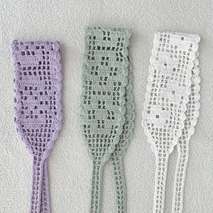 Puede incluir: Tres art&iacute;culos de ganchillo hechos a mano en colores pastel. Los art&iacute;culos son de color lila claro, verde suave y blanco. Cada art&iacute;culo tiene una parte superior rectangular con un patr&oacute;n calado decorativo y lazos largos y estrechos.