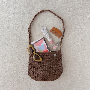 May include: A brown woven shoulder bag containing a white tube of Hello Berry hand cream, a small wooden comb, a clear glass perfume bottle, a pair of yellow cat-eye sunglasses, and a rectangular card featuring a colorful illustration of a person.