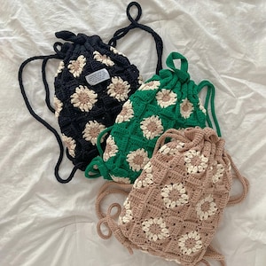 Puede incluir: Tres bolsas de cordón de ganchillo con diseños de flores de margarita. Las bolsas son negras, verdes y marrones.