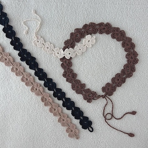 Può includere: Accessori floreali all'uncinetto in vari colori. L'immagine presenta una fascia per capelli all'uncinetto marrone, crema e nera, insieme a finiture floreali abbinate in nero e beige. Gli articoli fatti a mano sono esposti su uno sfondo neutro.