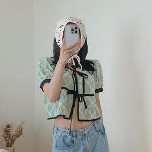 May include: A light green lace top with black trim and a bow closure. The top has short, puffed sleeves and is paired with light wash denim jeans. A white crocheted bonnet is also visible.