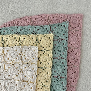 May include: A stack of four crocheted squares in varying pastel colors: white, pale yellow, mint green, and dusty rose. Each square features a floral pattern and a scalloped edge, showcasing a handmade craft.